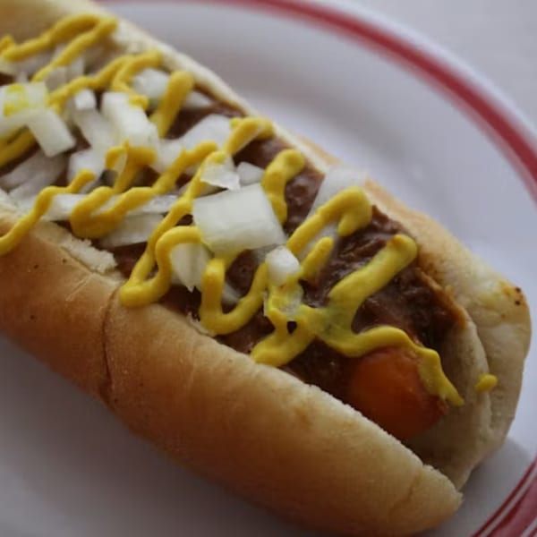 A close-up of a Coney Island hot dog with chili, diced onions, and mustard on a white plate.