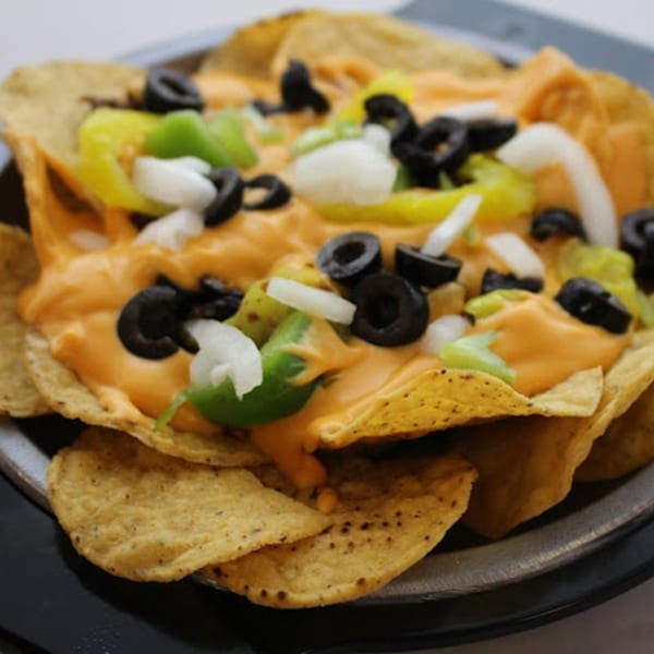 Nachos with melted cheese, black olives, green peppers, and white onions in a metal bowl.