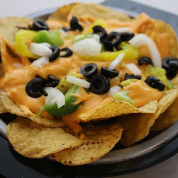 Nachos with melted cheese, black olives, green peppers, and onions on a dark plate.
